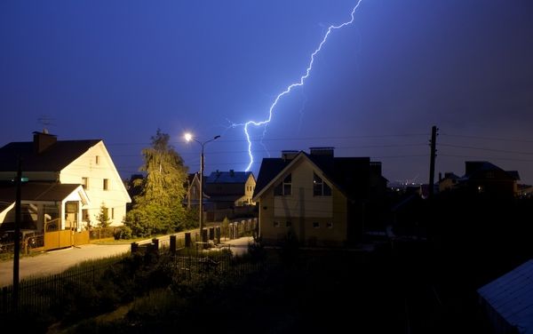 Ein Blitz schlägt in den Himmel über einem Wohngebiet mit mehreren Häusern. Eine Straßenlaterne leuchtet, Bäume und Zäune sind sichtbar.