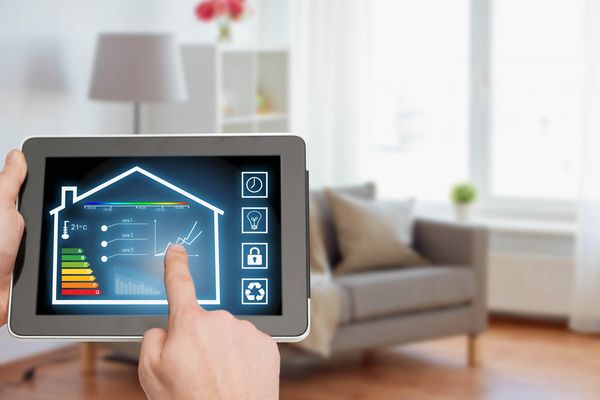Eine Hand zeigt auf ein Tablet mit einer grafischen Darstellung eines Hauses, das Energieverbrauchsdaten anzeigt. Im Hintergrund ist ein Wohnzimmer mit einem grauen Sofa und einem Regal zu sehen.