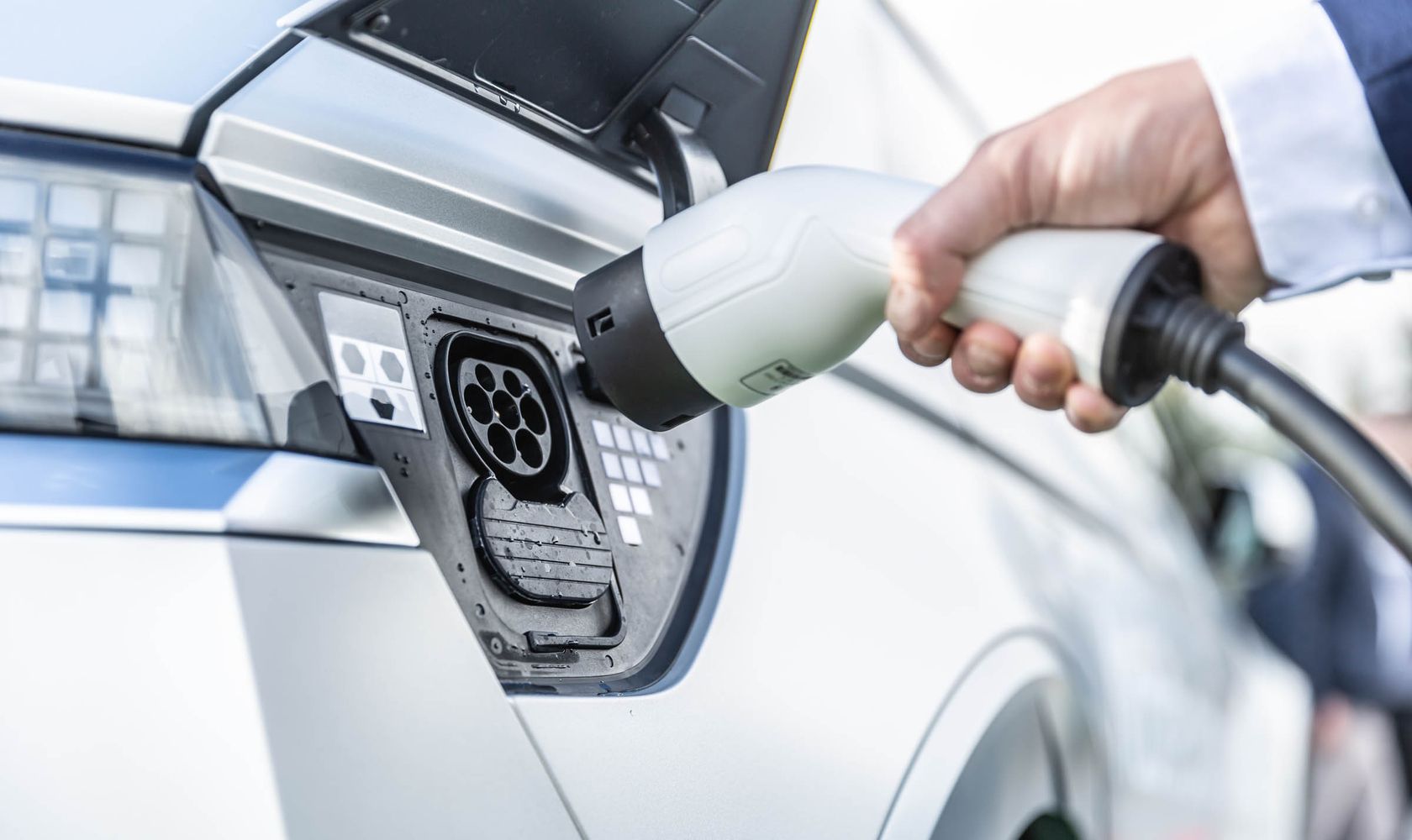 Eine Hand hält einen Ladestecker, der in die Ladebuchse eines Elektroautos eingesteckt wird. Das Auto ist silberfarben.