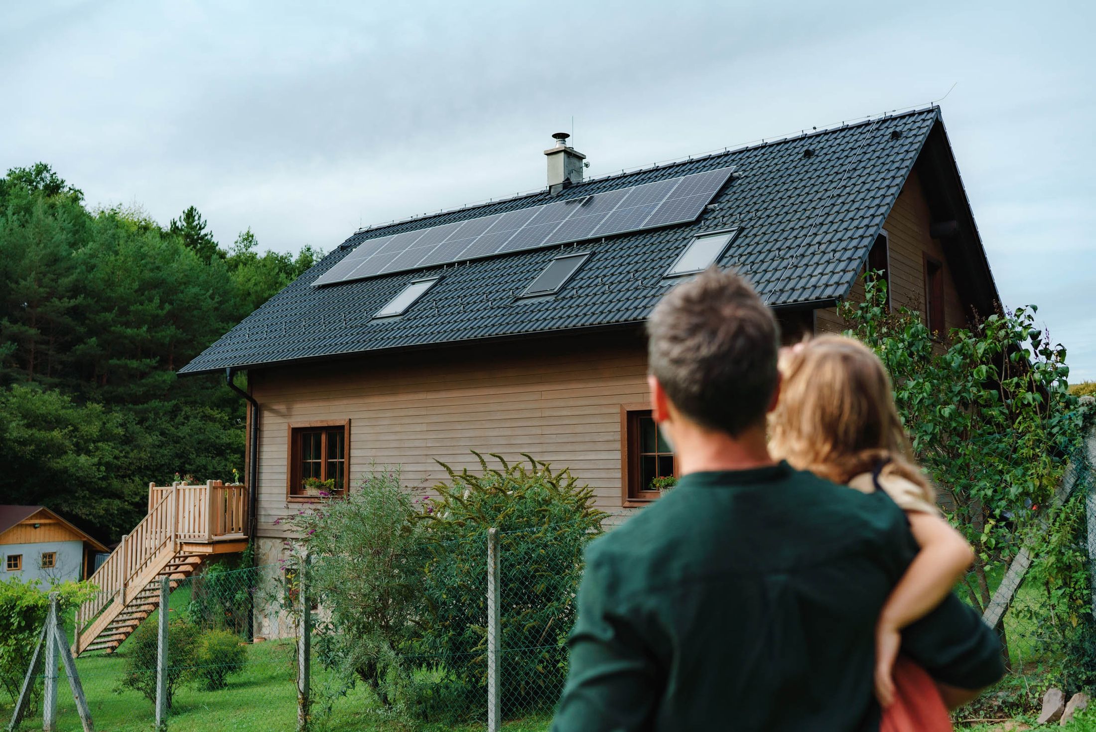 Ein Mann hält ein Kind auf dem Arm und blickt auf ein Holzhaus mit einem schwarzen Dach, das mit Solarpanelen ausgestattet ist. Im Hintergrund sind Bäume und ein Zaun sichtbar.